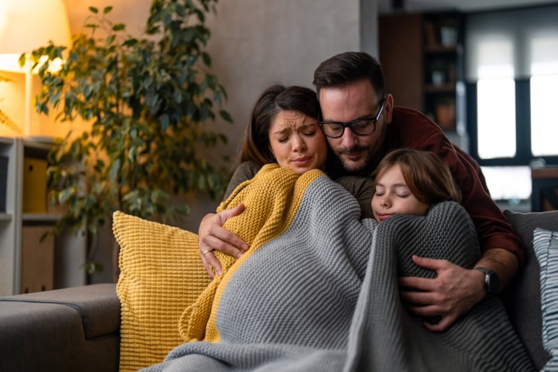 Why Isn’t My Furnace Heating My Entire Home? Photo of a family of three, freezing in their home, huddled under blankets to keep warm.