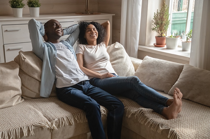 Air Filters vs. Air Cleaners. Man and woman couple happily resting on a beige couch with their feet up.