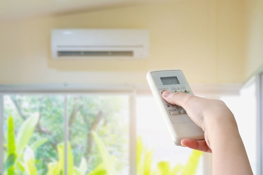 Planning to Remodel? Go Ductless! Image si a photograph of a ductless mini split mounted on a beige wall above a window out of focus. In focus, there is a hand holding a remote that is aimed at the mini split.